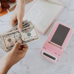 A person holding a pink cell phone next to a pile of payday loans money